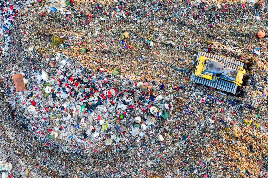 An aerial view of a large landfill site filled with a dense layer of mixed waste, including plastic bottles, paper, metal cans, and other household debris, creating a multicolored and textured surface. In the middle right of the image, a yellow tracked excavator with a large bucket attachment is actively engaged in compacting or sorting the waste, positioned on a dirt-covered area. The surrounding environment features uneven terrain with visible soil and scattered refuse, with no vegetation or natural landscape in sight. The scene is illuminated by natural daylight, highlighting the various materials' colors and textures, emphasizing the extent of rubbish accumulation characteristic of waste disposal sites. This image illustrates the scale and density of landfill operations, aligning with topics related to private waste handling or alternative rubbish collection methods employed by waste disposal companies such as Waste Disposal West Hampstead, operating within the context of independent rubbish removal services.