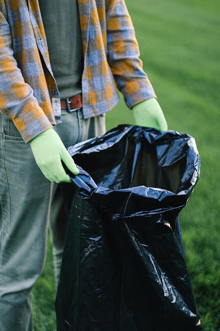 A person wearing a yellow and grey checkered shirt, grey trousers, and light green rubber gloves is holding open a large black garbage bag made of durable plastic material, standing outdoors on a grassy area. The bag is empty, with its top folded over, revealing the shiny black interior. The background consists of a well-maintained green lawn, and the scene appears to be part of a waste clearance activity, possibly related to rubbish removal or garden waste disposal. The individual’s hands are securely gripping the edges of the bag, ready for collecting rubbish, indicating a practical and organized approach to waste handling, often associated with private waste disposal services instead of municipal collection methods. The natural outdoor setting provides ample lighting, highlighting the textures of the gloves, bag, and the grass surrounding them. This scene emphasizes the process of independent waste management, aligning with services like those offered by Waste Disposal West Hampstead, specialising in rubbish removal.