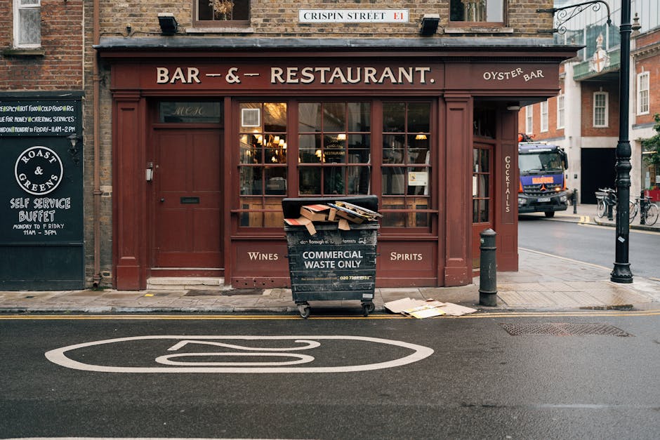 The image depicts the façade of a closed restaurant with a maroon wooden exterior, large glass windows, and a sign indicating it is a bar and oyster restaurant. In front of the establishment, a black commercial waste bin labeled 'COMMERCIAL WASTE ONLY' is placed on the sidewalk, filled with discarded papers and cardboard boxes that are spilling over the top. The bin is aligned near a small grey bollard and a black lamppost, both positioned on the pavement. Adjacent to the bin, there is a slightly crumpled piece of paper on the ground. On the left side, a black signboard advertising a self-service buffet runs along the sidewalk, and on the right, a parked truck is visible in the background. The scene is illuminated by natural daylight, and the street features typical urban elements such as a no-parking road marking, curbstones, and nearby bicycles, suggesting a city environment that handles various waste types, including commercial waste that might require private collection services like those provided by Waste Disposal West Hampstead.
