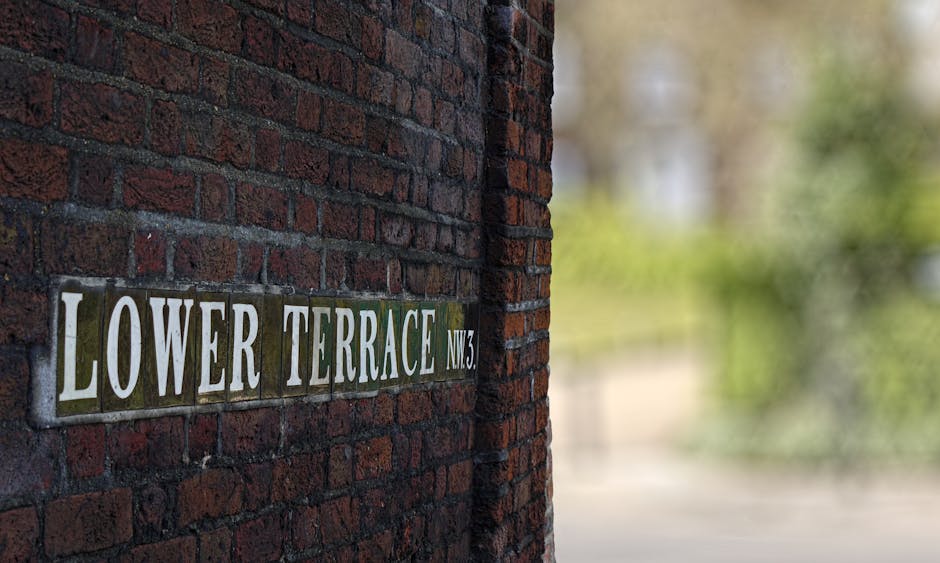 A close-up view of a street sign affixed to a dark red brick wall, displaying the name 'Lower Terrace NW3' in bold white and green letters. The brickwork has a rough, textured surface with visible mortar joints. The sign is made of ceramic tiles, with some ceramic edges slightly chipped, and the background of the sign is a muted green color. The wall appears to be part of a building or boundary wall on a residential street. In the background, the image blurs into a natural, leafy environment with hints of greenery and soft daylight, suggesting an outdoor setting. This scene is associated with an area where private rubbish collection or on-site clearance services might be relevant, reflecting the context of waste disposal and property maintenance in residential neighborhoods.