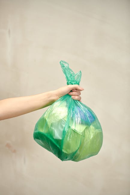 A person's hand is holding a green plastic rubbish bag tied at the top, with visible folds and creases and a slightly shiny surface. The bag appears full, containing assorted waste materials, and is suspended in mid-air against a plain beige wall background. The hand is positioned on the left side of the image, gripping the gathered top of the bag with fingers wrapped around it, with the arm extending from the left edge of the frame. The scene suggests an act of rubbish collection or disposal, relevant to services provided by waste disposal companies such as Waste Disposal West Hampstead, which handles independent waste removal for local flats and property complexes through various methods including on-site clearance or private collection solutions. The lighting is soft and even, illuminating the bag and hand clearly, with no shadows or additional objects in view, emphasizing the focus on waste handling procedures in residential or commercial environments.