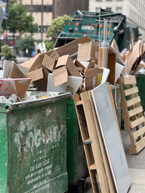 A large green rubbish bin filled with various discarded cardboard boxes of different sizes and shapes, some flattened and some partially assembled, along with a white polystyrene foam container. The textured surfaces of the cardboard display brown and beige tones, with some printed labels visible. The bin is situated outdoors on a city street, with a backdrop of a waste collection vehicle in dark green, partially obscured, and concrete buildings surrounding the area. To the right of the bin, there are wooden pallets leaning against it, and the scene is illuminated by natural daylight, highlighting the contrast between the clutter and the urban environment. This setting illustrates a typical scene of landfill-bound waste awaiting collection, relevant to private disposal services offered by Waste Disposal West Hampstead for alternative rubbish removal options in NW6.