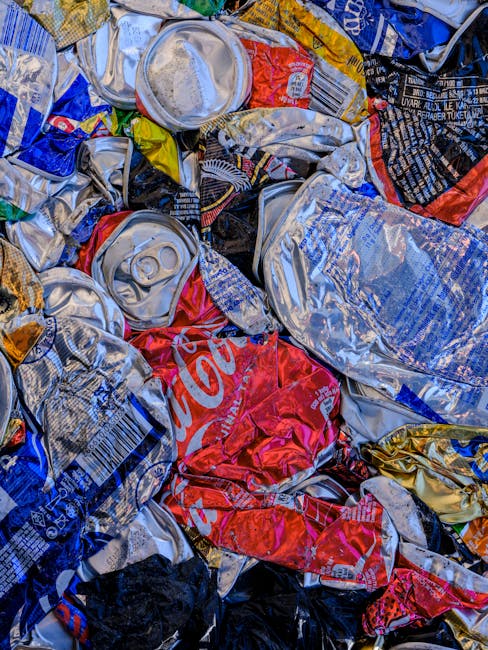 A close-up view of a pile of crushed aluminum beverage cans, predominantly silver with some red, blue, black, and yellow labels. The cans display varied textures, from shiny and smooth to slightly dented and crumpled, with visible pull-tabs and printed branding, including a partial Coca-Cola logo. The cans are stacked haphazardly, overlapping each other, with some partially flattened. The background is not distinguishable, but the cans appear to be on a flat surface, possibly prepared for collection or disposal. This scene reflects an aspect of waste management involving the collection of recyclable aluminum cans, often handled by independent waste disposal services like Waste Disposal West Hampstead, which supports alternative methods of rubbish removal outside standard municipal collections.