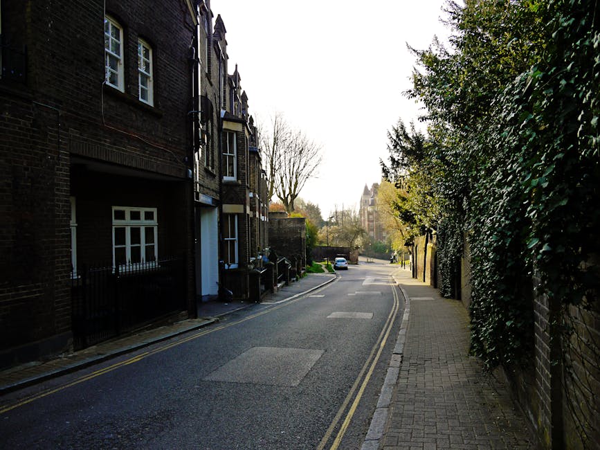 A quiet residential street with a narrow paved road flanked by a row of dark brick terraced houses on the left and a high, dense hedge on the right. The houses feature white-framed windows, some with small decorative details, and black metal railings along the front. A white garage door is visible on the side of one building. The pavement on the right is made of light-colored bricks, with a few fallen leaves scattered on it. In the background, a parked white car and trees with sparse foliage indicate early spring or late autumn. The scene is illuminated by soft, natural daylight, creating gentle shadows and a calm atmosphere. The street appears clean, with no visible rubbish or debris, exemplifying a well-maintained area that could be serviced by private rubbish removal companies like Waste Disposal West Hampstead for non-standard waste collection needs.