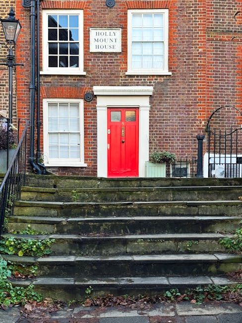 The image depicts the front façade of a brick residential building with a red wooden door featuring small glass panes at the top, surrounded by a white frame. To the left of the door, there are three wide, white-paneled windows with closed blinds; two are positioned on the ground floor, and one is on the upper level, directly above the door. The brickwork is a traditional red hue with some darker accents, and the building has a white sign on the wall reading 'HOLLY MOUNT HOUSE'. In front of the building, there is a short flight of moss-covered stone steps leading to a small landing area, flanked on both sides by black metal railings. On the right side of the steps, there is a small rectangular planter with a bushy green plant, and another larger light green planter container nearby. To the left, a black lamppost stands adjacent to the staircase, and the surrounding environment includes small patches of greenery growing between the steps and a bit of fallen leaves at the base, suggesting an outdoor setting. This scene captures the exterior of a traditional residential property, illustrating a typical urban or suburban street view consistent with private property standards often associated with independent waste collection or on-site clearance options, relevant to waste disposal services such as those by Waste Disposal West Hampstead.