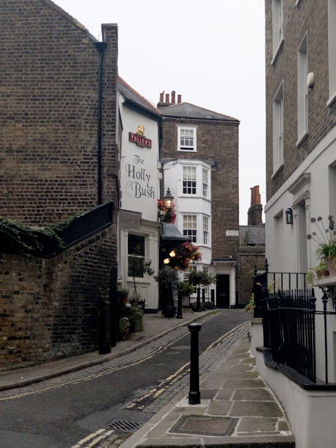 A narrow urban street scene in West Hampstead featuring a small, curved cobblestone road bordered by brick and white-painted buildings. On the left, an older brick wall partially covered with greenery and a small outdoor area with potted plants and hanging flower baskets along the building facade. In the middle distance, a traditional pub sign for 'The Holly Bush' is visible, with adjacent hanging flower baskets adding color and texture. The buildings have multiple stories with sash windows, some with decorative molding, and brick chimneys on rooftops. On the right, a white building with a small balcony adorned with potted plants and a black metal railing. The scene appears to be during daylight, with overcast sky casting diffuse lighting over the scene, suggesting an environment that may be suitable for private waste handling or alternative rubbish collection, often requiring on-site or independent disposal options. Waste Disposal West Hampstead specializes in managing such waste removal within urban residential settings, ensuring clutter is cleared efficiently from similar narrow streets and alleyways.